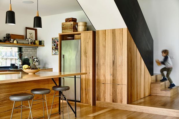 Warm open-plan kitchen with a timber island and three stools, exposed shelving, brick-tile splashback, stainless-steel refrigerator, and extensive vertical-grain timber cabinetry. A bold dark diagonal stair structure frames the right side, leading to a raised timber-floored area. Black pendant lights and timber flooring add contrast.