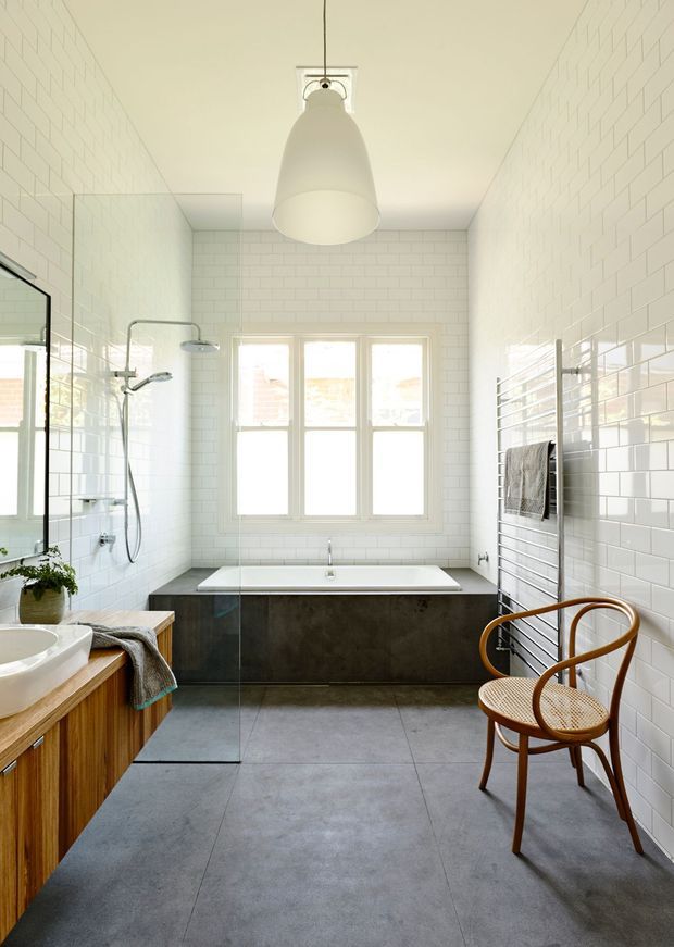 Bright contemporary bathroom with white subway-tiled walls, a freestanding bath set against dark cabinetry or a dark tiled base, large grey floor tiles, a frameless glass shower enclosure, timber vanity cabinetry, and a chrome heated towel rail. A large white pendant light hangs centrally, while a cane and timber chair adds warmth beside the bath. Natural light enters through the white-framed rear window.