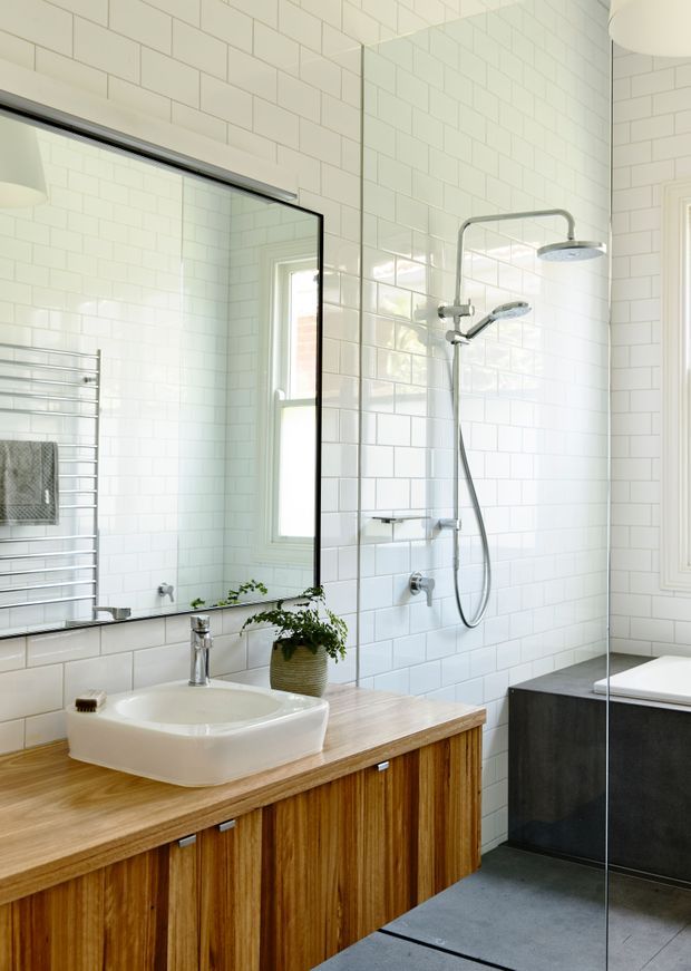 Bright contemporary bathroom with white subway-tile walls, a large black-edged mirror, timber vanity with a white vessel basin, chrome tapware, a wall-mounted towel rail, and a frameless glass shower enclosure. A dark tiled bath surround is visible at right. Natural light enters through tall windows, creating a clean, airy interior.