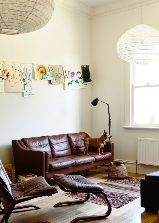 Bright, eclectic living room with tall cream walls, a large window, timber flooring, a worn brown leather sofa, matching leather lounge chairs and ottomans, a black adjustable floor lamp, patterned area rug, woven basket, and colourful children's artwork displayed along the wall. Two oversized woven pendant lights hang from the ceiling, and a small dog is standing on the sofa.