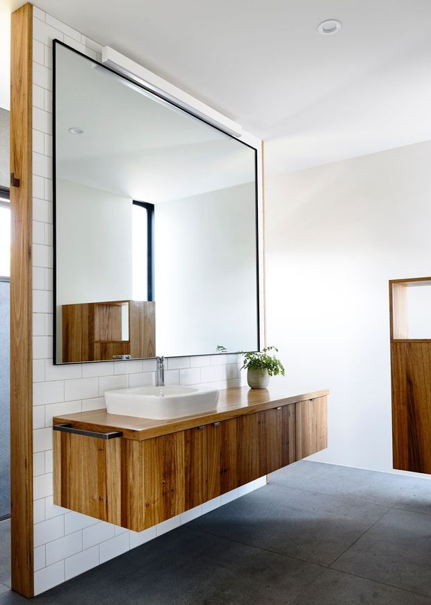 Bright contemporary bathroom featuring a wall-mounted timber vanity with integrated storage, a white vessel basin, chrome tapware, a large frameless-edged mirror, and white rectangular wall tiles. The room has a dark grey tiled floor, recessed ceiling lighting, and a timber-framed doorway.