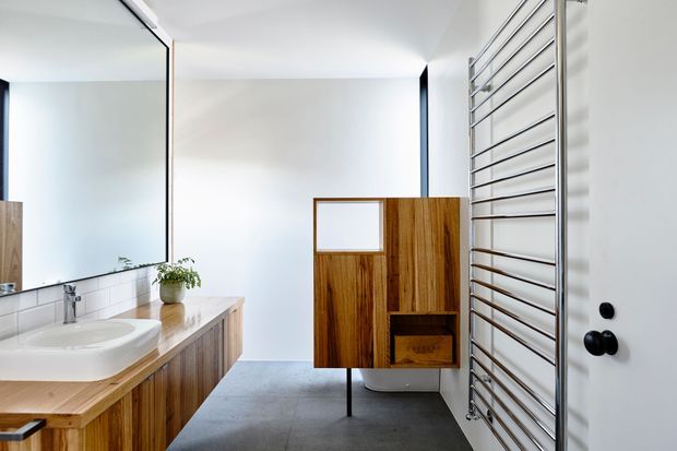 Bright contemporary bathroom featuring a wall-mounted white basin on a timber vanity, white tiled walls, a large frameless mirror, grey concrete-look flooring, a freestanding timber storage cabinet, and a tall chrome heated towel rail. Minimal black hardware adds contrast.