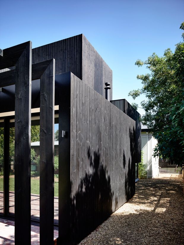 Exterior view of a contemporary dark timber-clad building or extension, featuring bold vertical timber screens and projecting wall planes. The blackened vertical cladding and substantial timber posts create a strong geometric facade, with a gravel-covered outdoor area, mature planting, and glimpses of glazing and an adjacent light-coloured building.