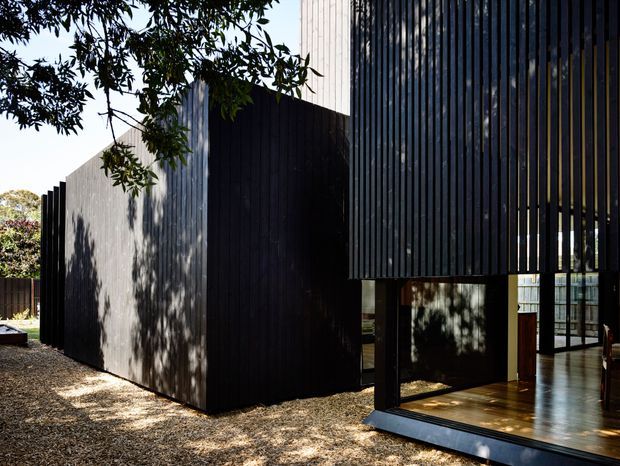 Exterior view of a contemporary black-clad building with strong vertical timber screening and elevated glazed living spaces. The design uses simple rectilinear volumes, a partially open ground level, and warm timber flooring visible through the glazing. Trees and gravel landscaping frame the structure.