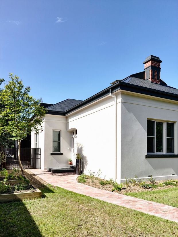Exterior view of a single-storey rendered home with a dark hipped roof and prominent red brick chimney. A pale brick-paved walkway leads to the recessed entry, with a small landscaped garden, raised planter beds, lawn, and dark vertical fencing visible around the property.