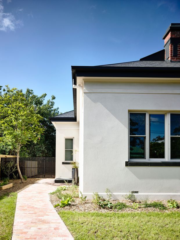 Exterior side view of a refined rendered residential building with a dark hipped roof, projecting cornice, white-framed windows, and a prominent brick chimney. A red brick garden path leads alongside landscaped planting and a timber boundary fence.