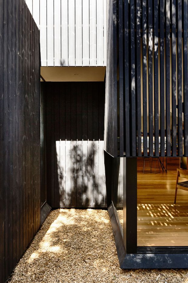A narrow, modern architectural exterior passage or courtyard framed by dark vertical timber cladding and slatted screening. White vertical cladding forms an upper volume, while a large glazed opening reveals a warm timber-floored interior with simple seating. Strong sunlight creates dappled shadows across the pale wood-chip ground and facade surfaces.
