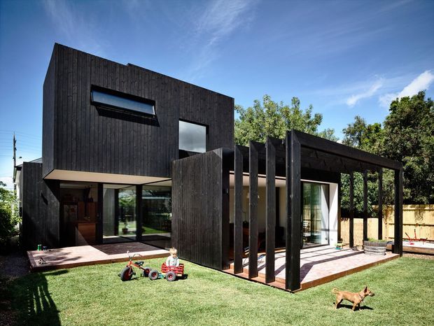 Exterior view of a contemporary dark-clad two-storey house with a projecting upper volume, large glazed openings, and a covered outdoor terrace or pergola extending into a lawned backyard. The facade uses vertically oriented black timber cladding, with a timber deck, mature trees, and a high boundary fence visible around the property.
