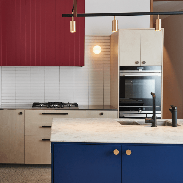 A contemporary kitchen featuring a deep blue island with a pale stone benchtop, light timber cabinetry, a white horizontal-tile splashback, integrated black appliances, and a bold burgundy-red wall panel. Black linear track lighting with brass cylindrical pendants and a dark sculptural tap add contrast.