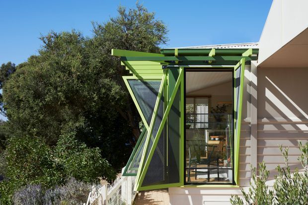 Exterior view of a contemporary residential building with a distinctive bright green louvred and framed sun-shading structure around a large glazed opening. The projecting screen includes angled supports and operable-looking panels, casting strong linear shadows across the pale rendered facade. Interior furnishings are visible through the glazing, while mature vegetation surrounds the side of the building.