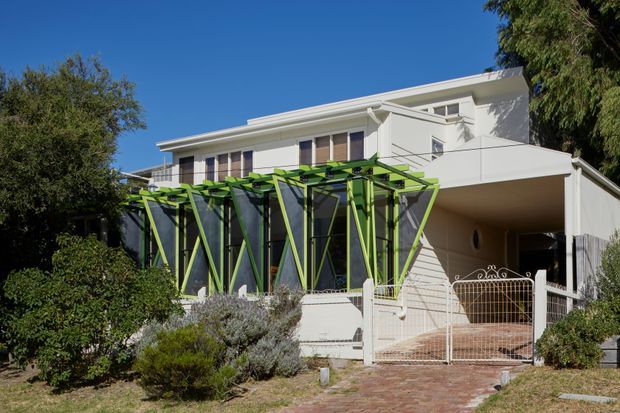 Exterior view of a two-storey white rendered residence with a flat or very low-pitched roof, projecting upper-level volume, and a distinctive lime-green and dark structural pergola or sunshade over the glazed ground-floor facade. The front garden includes established shrubs, a brick-paved path, and a metal pedestrian gate. Bright clear daylight emphasizes the strong contrast between the white building envelope and vivid green architectural screening.