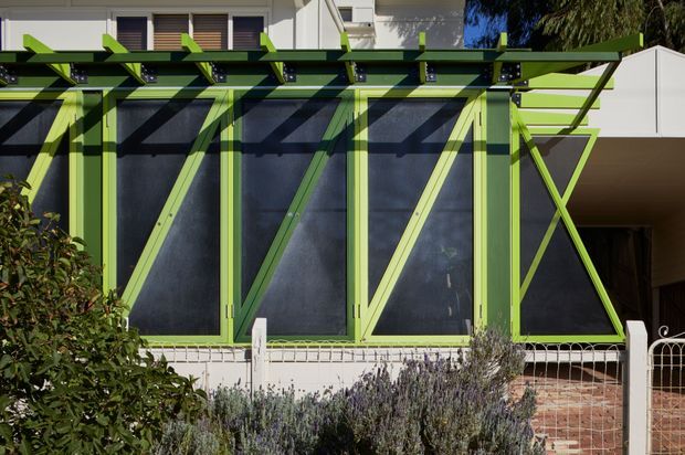 Exterior view of a contemporary residential facade featuring a long glazed wall or sunroom enclosed by vivid lime-green timber framing and diagonal bracing. A projecting pergola with green beams shades the glazing, while a low white wall, brick paving, planting, and a simple metal fence occupy the foreground. The image emphasizes the bold structural frame and screened glazing.