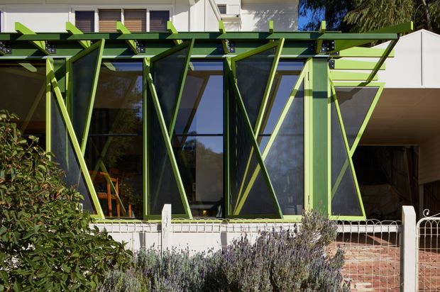Exterior view of a contemporary residential facade featuring a dramatic lime-green structural sunshade or pergola frame across full-height glazing. Dark green diagonal bracing creates a bold geometric pattern over the windows, with white upper wall surfaces, a brick-paved entry area, and landscaped planting in the foreground.