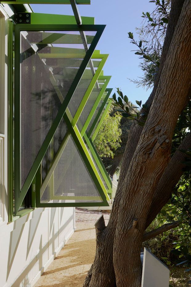 Exterior architectural detail showing a row of projecting green-framed window sunshades or awnings with perforated metal screening along a light-coloured building. A mature tree runs beside the facade, and strong sunlight creates crisp shadows on the wall and walkway.