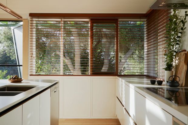 Bright contemporary kitchen with extensive timber-framed windows and horizontal timber blinds overlooking leafy trees. White paneled cabinetry wraps around the room, with dark stone or composite benchtops, a stainless-steel sink, integrated dishwasher, timber flooring, and a red-brown brick feature wall. Natural light creates strong striped shadows across the cabinetry and work surfaces.
