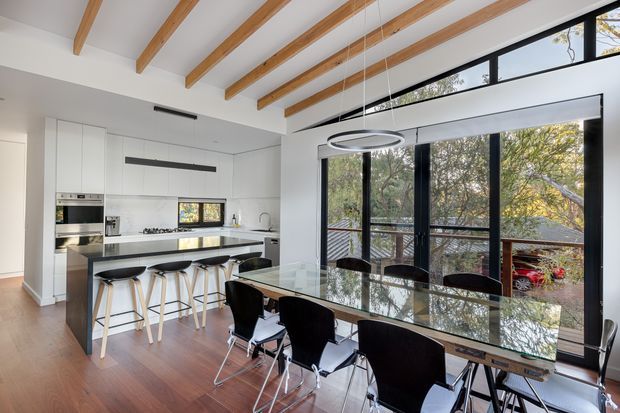 Bright open-plan kitchen and dining area with white flat-panel cabinetry, a long island with bar seating, integrated stainless-steel appliances, and a glass dining table with black chairs. The room features warm timber flooring, exposed timber ceiling beams, large black-framed windows and doors, and a modern circular pendant light overlooking a leafy outdoor setting.