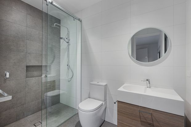 A contemporary bathroom with a frameless glass walk-in shower, grey tiled shower walls and floor, a white wall-mounted toilet, and a floating timber-look vanity with an integrated white basin. Large white wall tiles and a round mirror create a clean, bright, minimalist look.