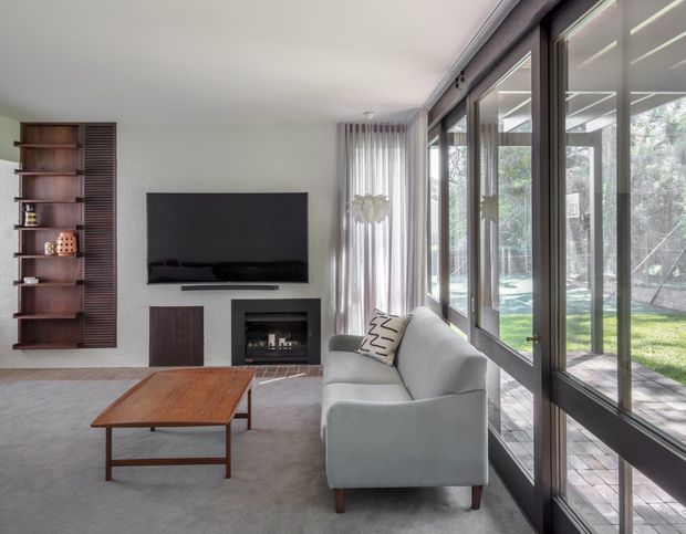 A bright contemporary living room with a large grey sectional sofa facing a wall-mounted television and a built-in fireplace. Floor-to-ceiling glazed doors and windows line one side of the room, opening views to a timber deck and landscaped backyard. The room also features a warm timber coffee table, a dark timber open shelving unit, neutral carpeting, sheer curtains, and dark-framed glazing.