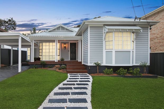 Front exterior view of a single-storey heritage-style home with a light grey weatherboard facade, prominent gabled roof forms, enclosed front verandah, decorative bay window, covered carport, and a landscaped lawn with a stepping-stone path.