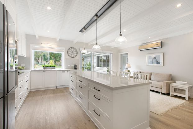 Bright open-plan kitchen and living area with white shaker-style cabinetry, a large central island, pale timber-look flooring, white panelled ceiling, pendant lights, dining seating, and a compact lounge area. Large windows bring in greenery and abundant natural light.