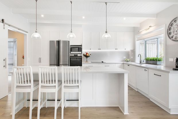 Bright white contemporary kitchen with a central island and integrated breakfast seating. The room features white Shaker-style cabinetry, stainless-steel refrigerator and appliances, a pale reflective splashback, pendant lights, light timber-look flooring, and a large window over the benchtop. A dining area and adjoining room are visible through the doorway.