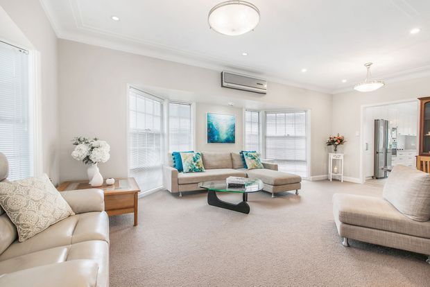 Bright, spacious living room with pale neutral walls, white trim, carpeted flooring, several upholstered sofas and armchairs, a glass coffee table, bay-style windows with blinds, recessed lighting, and a central flush-mount ceiling fixture. A compact kitchen or utility area is visible through the opening at the rear.