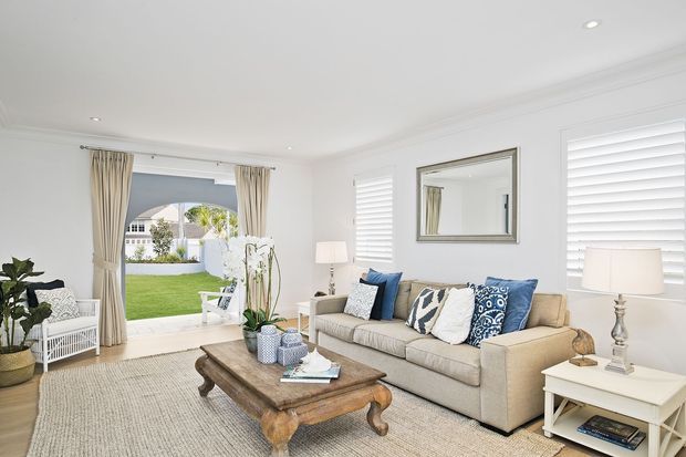 Bright, traditionally styled living room with white walls, recessed ceiling lights, plantation shutters, full-height curtains, and a neutral upholstered sofa arranged around a substantial carved timber coffee table. A large opening leads to a manicured lawn and garden outside, while decorative cushions and indoor plants add blue and green accents.