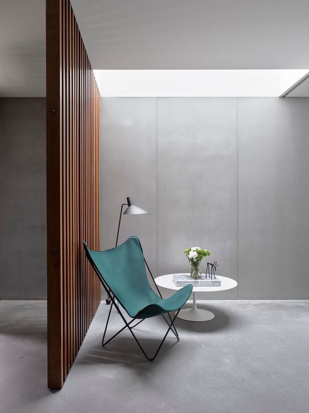 A minimalist interior vignette with a teal butterfly chair, slim black floor lamp, and round white pedestal table against smooth grey concrete walls and floor. A vertical dark timber slatted screen divides the space, while a long overhead skylight introduces soft natural light. The composition is sparse, sculptural, and contemporary.