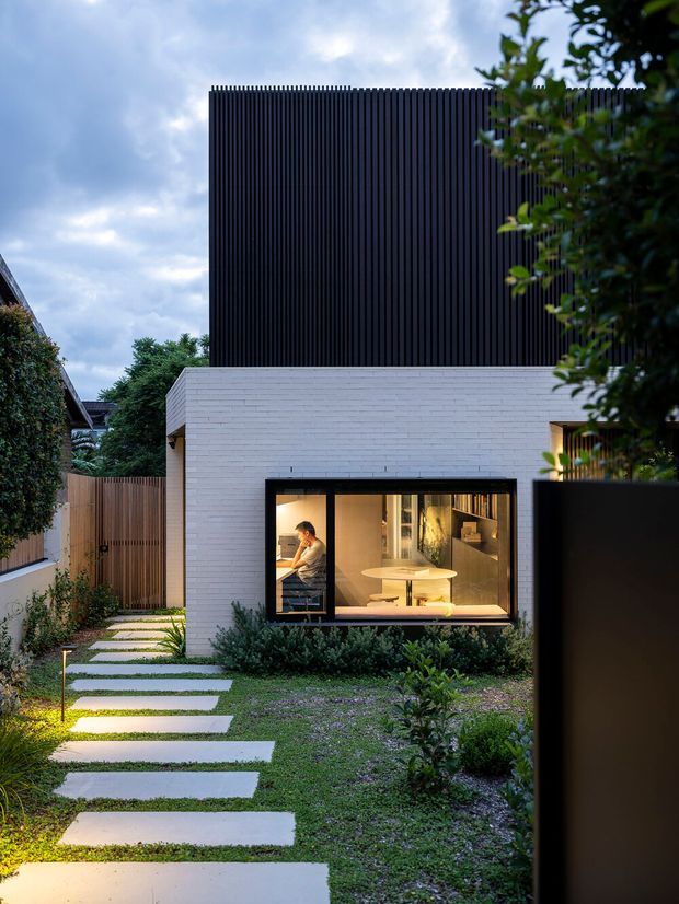 Exterior view of a contemporary two-storey residence at dusk, featuring a white brick lower level with a large black-framed window and a tall upper volume clad in closely spaced dark vertical battens. A landscaped front garden includes a stepping-stone path, lawn, planting, timber screening and subtle pathway lighting. Warm interior lighting reveals a compact living or dining area through the window.