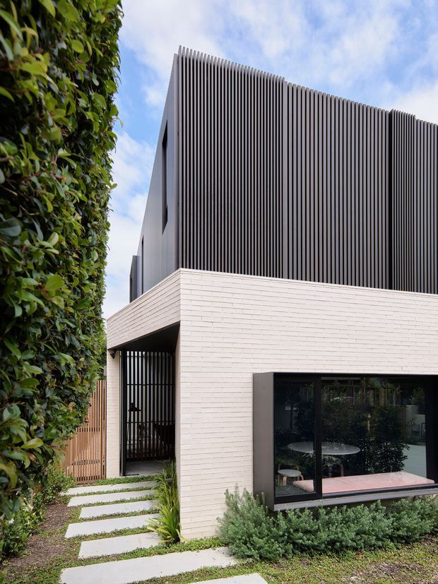 Contemporary multi-level residential facade viewed from a landscaped front path. The lower volume is clad in pale horizontal brick, with a deep dark-framed glazed opening and a recessed entry screened by vertical timber slats. The upper storey is wrapped in closely spaced dark vertical fins, creating a strong textured privacy facade. Dense hedging, low planting, and concrete stepping stones soften the minimalist architecture.