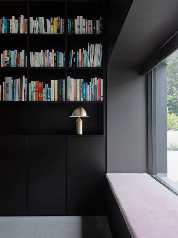 A dark, refined reading nook with a floor-to-ceiling black built-in bookcase filled with books and lower closed cabinetry. A metallic dome table lamp sits on the shelving, while a soft upholstered window seat runs along large glazed openings. The space uses strong black and charcoal tones with natural light entering from the side, creating a moody contemporary interior.