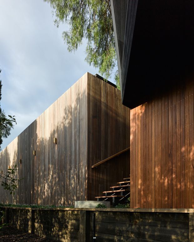Exterior architectural view of a contemporary timber-clad building composed of angular, box-like volumes. Vertical weathered and warm-toned timber boards form the facades, with a recessed entrance or stair opening between the volumes. The building is elevated above a dark retaining wall or platform, with integrated wall lights and strong tree-shadow patterns across the cladding.