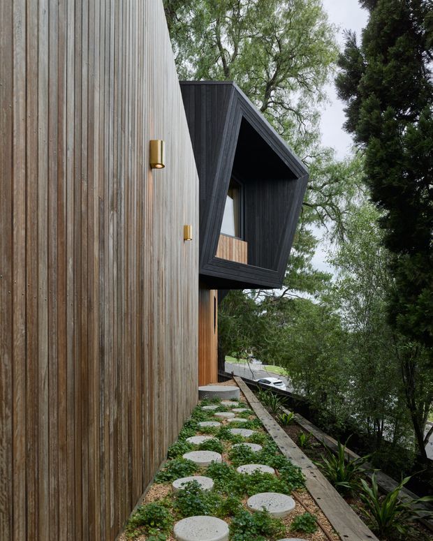 A contemporary residential exterior viewed along a narrow landscaped side path. The facade combines vertically aligned natural timber cladding with a projecting, dark-clad angular bay or balcony with deep reveals. Small brass-toned wall lights punctuate the timber wall, while stepping-stone pavers and planted beds lead toward the elevated structure amid mature trees.