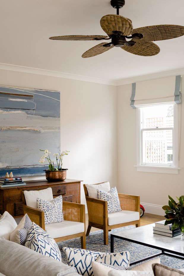 A bright, refined living room with cream walls, white trim, and a large abstract blue-grey artwork above a traditional wooden sideboard. A woven rattan ceiling fan forms a prominent feature overhead. Two wood-framed lounge chairs with white cushions and blue patterned pillows face a low black-framed coffee table, with part of a neutral sofa visible in the foreground. A window with a pale blue-grey Roman blind brings in daylight, while a patterned rug and indoor plants add warmth and texture.