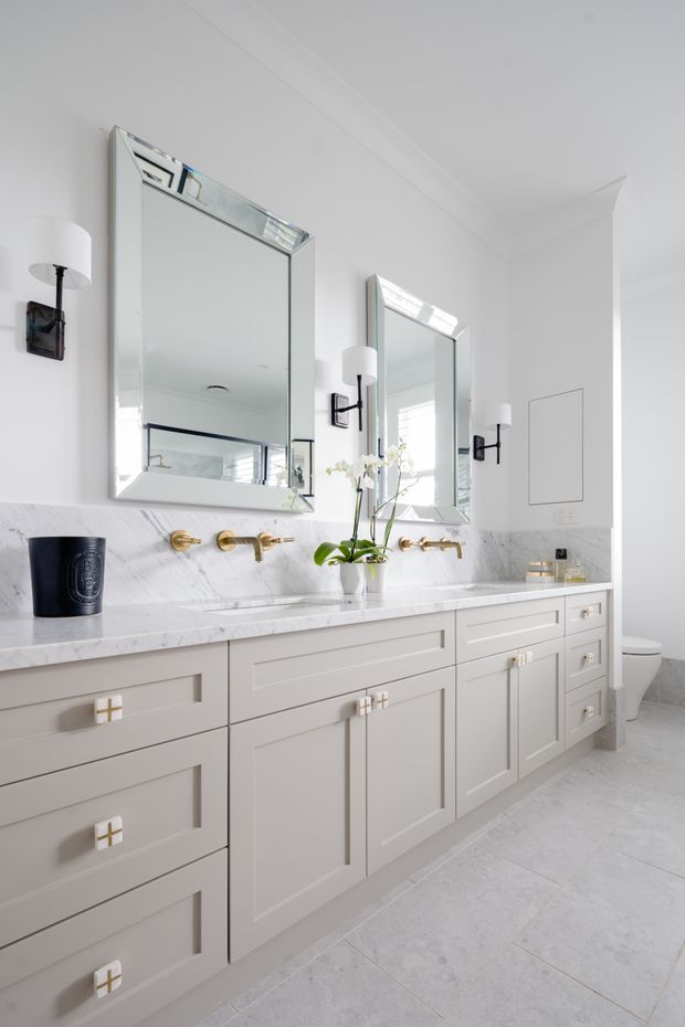 Bright, refined bathroom vanity with a long white built-in cabinet, paired rectangular mirrors, brass wall-mounted tapware, white marble-look countertop and backsplash, and black-and-white wall sconces. A toilet is partially visible through the opening on the right. The room has a clean, classic feel with abundant natural light and pale stone-look floor tiles.