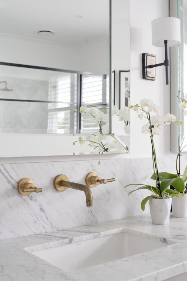 Bright, refined bathroom vanity featuring a white marble benchtop and splashback with integrated rectangular basin, wall-mounted aged brass tapware, a large mirrored cabinet, and a black-and-brass wall sconce. White orchids and plantation shutters add a fresh decorative touch.