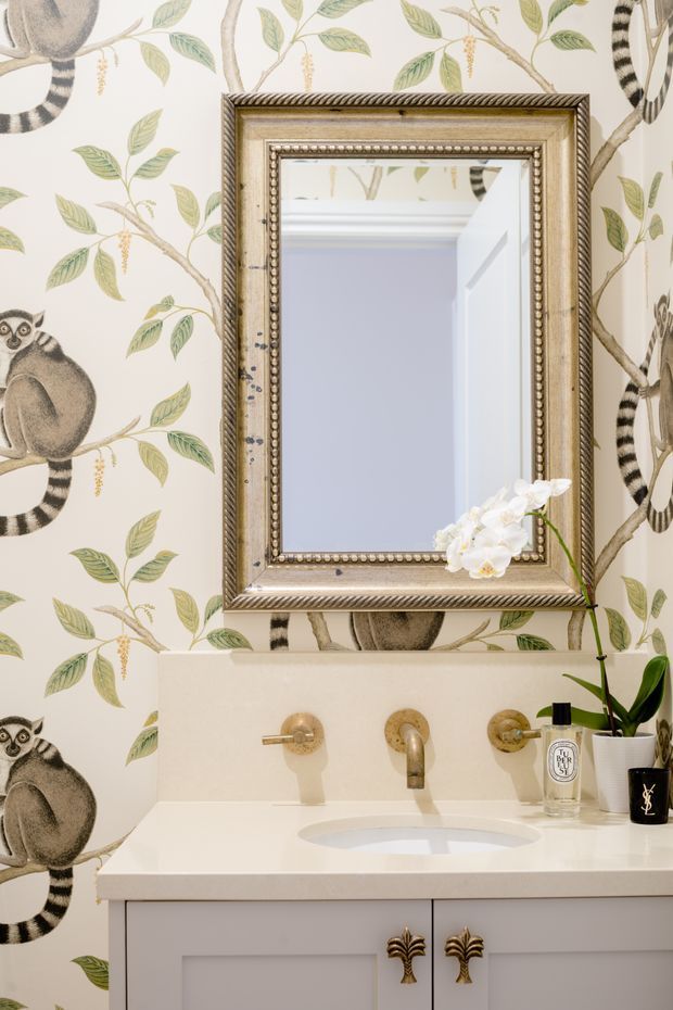 A refined bathroom vanity with a white integrated basin, pale cabinetry, and brushed brass wall-mounted tapware. The wall is covered in whimsical botanical and lemur-patterned wallpaper, while an ornate aged metallic-framed mirror hangs above the vanity. A white orchid and toiletries sit on the counter, creating a decorative, eclectic interior.