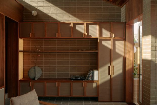 A warm, wood-lined living room storage wall with extensive built-in cabinetry, shelving, and a tall wardrobe. The joinery has dark timber framing and muted brown-grey inset panels, set against a small rectangular brown tile wall. Sunlight enters through the timber-framed glazing on the right, creating strong geometric shadows. A small portion of a sofa is visible in the foreground, along with books and decorative objects on the shelving. The space has a restrained mid-century character with rich natural materials and carefully composed built-in storage.