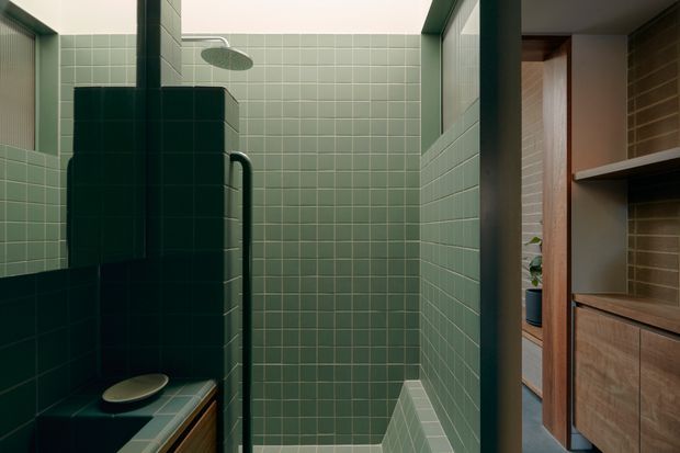 A compact, moody green-tiled bathroom focused on a walk-in shower. The shower and surrounding walls are clad in small square sage and deep green tiles, with a ceiling-mounted rain shower and dark metal grab rail. A tiled ledge with a shallow vessel is visible in the foreground, while warm timber bathroom cabinetry and open shelving appear to the right.