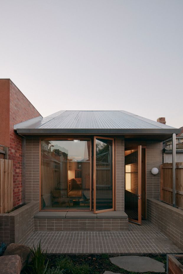 A compact rear courtyard extension viewed from outdoors, with a low-pitched corrugated metal roof, brown brick walls, large timber-framed glazed doors and windows, and a small paved brick terrace enclosed by timber fencing and brick privacy walls. Warm interior lighting is visible through the open door and glazing.