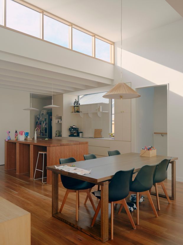 A bright, open-plan kitchen and dining area with a high ceiling and clerestory windows. The kitchen features a large timber island with white benchtops, integrated shelving, and appliances. A substantial timber dining table with dark teal upholstered chairs occupies the foreground. Exposed painted ceiling beams, warm timber flooring, minimalist white cabinetry, and pendant lights create a warm mid-century-influenced contemporary interior.