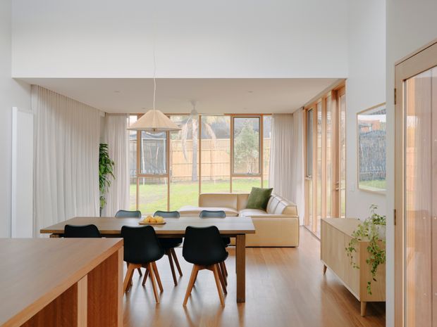 A bright open-plan interior combining a dining area and living room, with warm timber flooring, a large timber dining table, black dining chairs, a cream sofa, and extensive floor-to-ceiling glazing with sheer curtains. Light timber-framed doors and windows connect the room to a green backyard. Fixed timber cabinetry and a kitchen island or benchtop are visible in the foreground, while a pendant light hangs above the dining table.