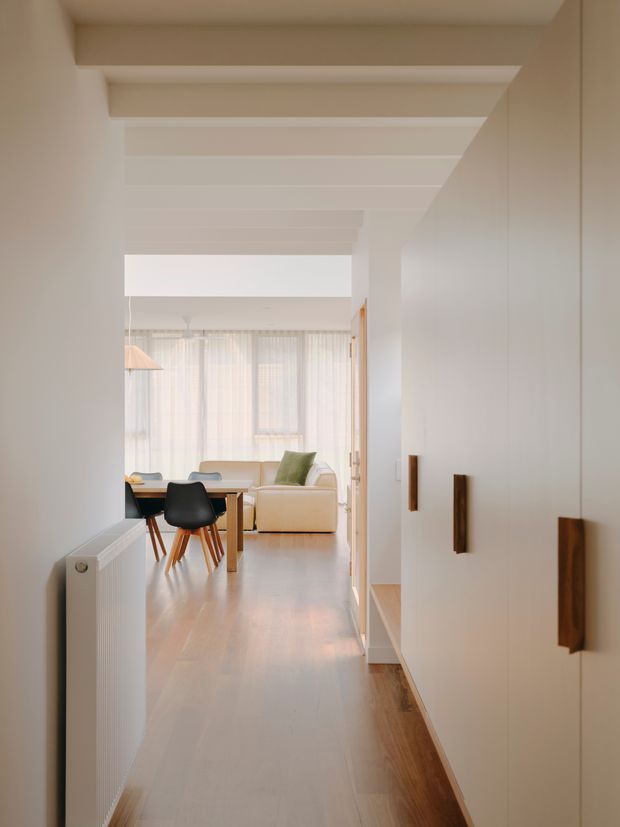 A bright, narrow residential hallway leading into an open dining and living area. The corridor has white walls, a white-painted exposed-beam ceiling, pale timber flooring, a white radiator, and tall built-in cabinetry with dark timber handles. In the background, a dining table with black chairs and a cream sofa sit beside softly filtered floor-to-ceiling windows. The interior is warm, minimal, and light-filled.