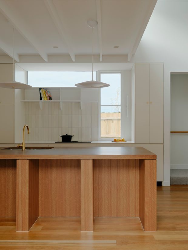 A warm, minimalist kitchen centred on a substantial timber island with open-sided supports and a smooth stone-look benchtop. The space features white vertical-tile splashback, white built-in cabinetry, timber flooring, exposed painted ceiling beams, pendant lights, and large windows bringing in soft natural light. A hallway and carpeted stair area are visible beyond the kitchen.