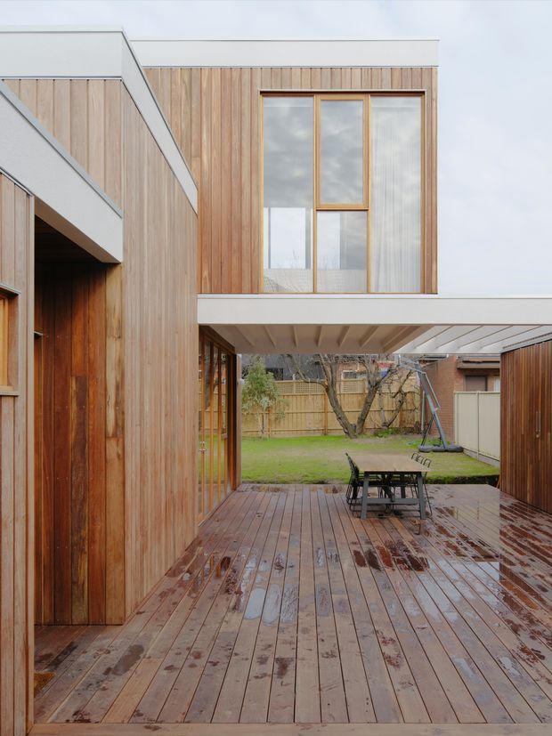 Exterior view of a contemporary two-storey timber-clad house organised around a covered outdoor deck or courtyard. Vertical natural timber cladding, white projecting box forms, large timber-framed windows, and an upper-level cantilever create a clean modern composition. The wet timber deck contains outdoor dining furniture and opens toward a fenced suburban garden.