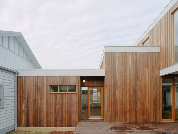 Exterior view of a contemporary residential building with warm vertical timber cladding, crisp white fascia and trim, and contrasting grey-white weatherboard walls. The composition shows connected low- and tall-volume forms, timber-framed glazing, a timber deck or paved entry terrace, and a simple flat-roofed link beneath an overcast sky.