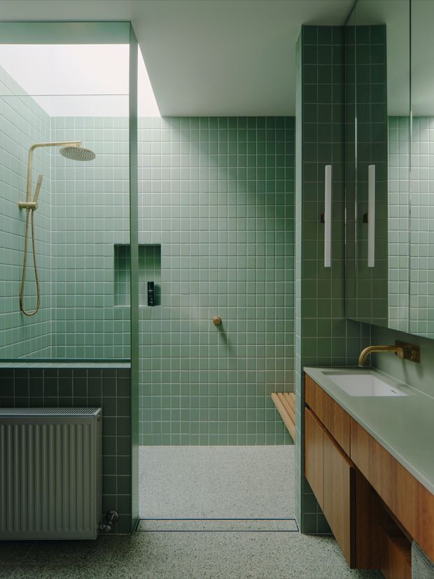 A compact contemporary bathroom with a walk-in shower, fully clad in small pale green square tiles. The shower has a frameless glass screen, exposed rainfall showerhead and brass fittings. A timber vanity with integrated white basin and brass wall-mounted tap occupies the right side, while a dark grey radiator sits beneath the window on the left. The floor is light terrazzo, and a large mirror reflects the tiled room.