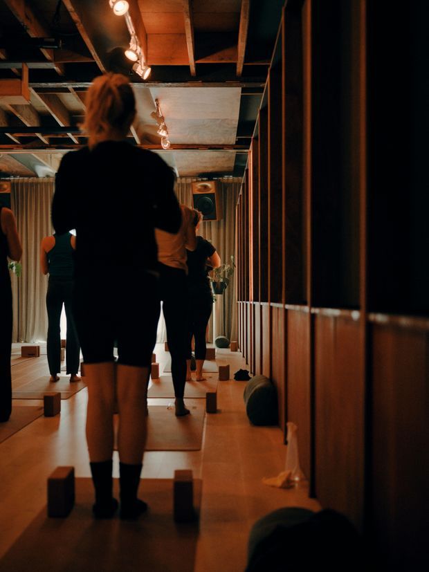 A dimly lit indoor fitness or yoga studio with several people exercising on individual mats. The space features warm timber flooring, extensive timber wall panelling and vertical slatted partitions, exposed ceiling beams and services, track lighting, and mirrors reflecting the room and participants. Yoga blocks and exercise equipment are distributed around the studio, creating a warm, atmospheric atmosphere.
