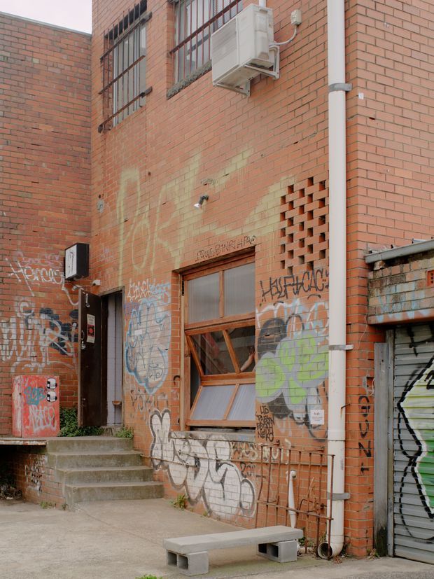 A close-up exterior view of a weathered red-brick urban building facade in a service laneway or courtyard. The wall features barred windows, a wall-mounted air-conditioning unit, exposed drainage pipe, a timber-framed window, a roller shutter, a small ventilation brick screen, graffiti, utility fixtures, concrete steps, and a simple concrete bench. The scene has a gritty adaptive-reuse or industrial urban character.