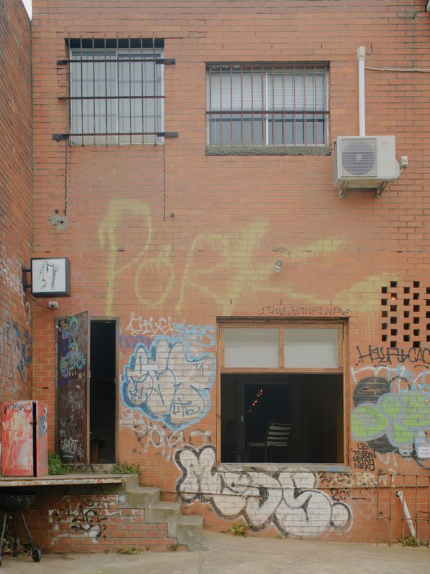 Exterior view of a red brick urban building facade with barred windows, a wall-mounted air-conditioning unit, graffiti-covered walls and doors, a small entrance stair, and a partially open ground-floor doorway. The scene has a gritty, heavily weathered streetscape character.